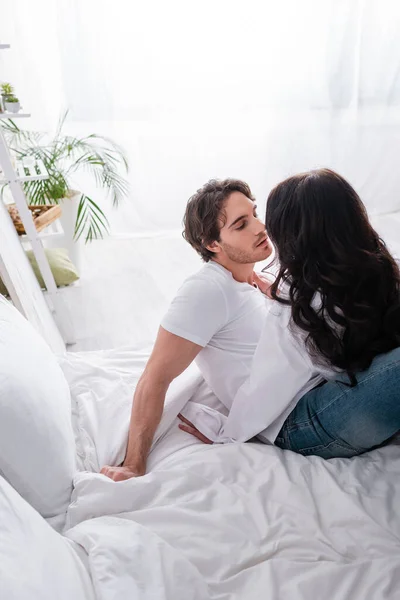 Брюнетка целует мужчину в белой футболке и джинсах в спальне — стоковое фото
