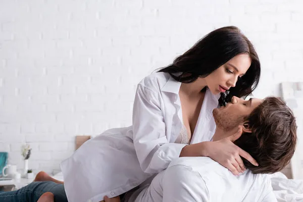 Passionate woman in unbuttoned shirt embracing young man in bedroom — Stock Photo