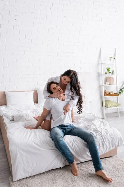 Пристрасна жінка обіймає чоловіка в джинсах, сидячи на ліжку з закритими очима — стокове фото