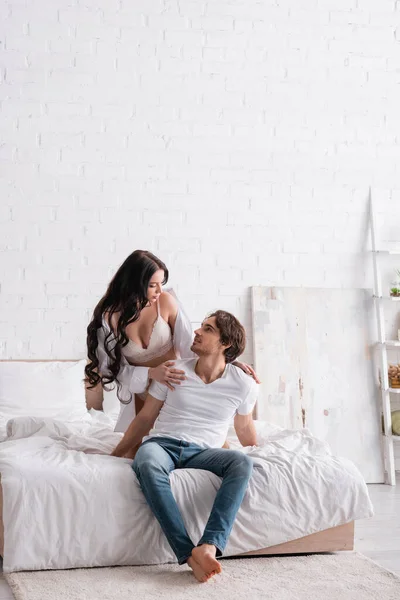 Сексуальна жінка в білій сорочці і бюстгальтер торкається плечей чоловіка, спокушаючи його в спальні — стокове фото