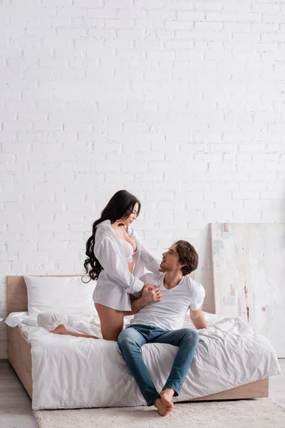 Mujer apasionada en camisa y sujetador seducir a hombre joven sentado en la cama en jeans - foto de stock