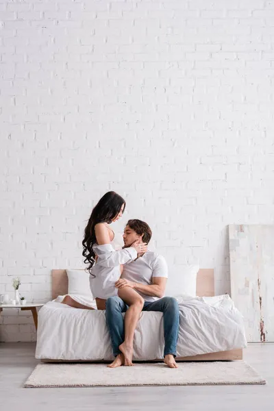 Woman in white shirt hugging boyfriend sitting on bed in jeans — Stock Photo