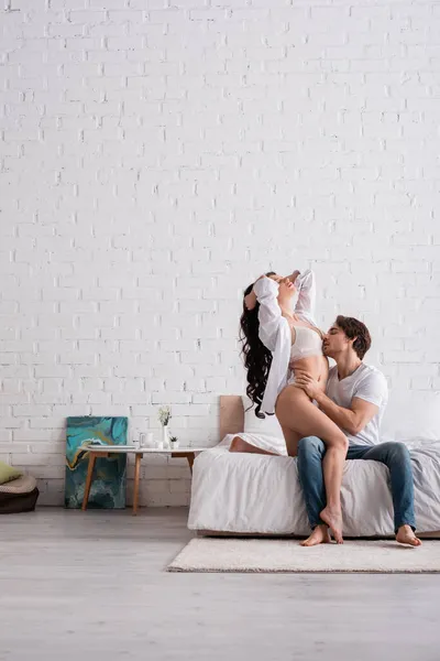 Man in jeans embracing passionate woman in lingerie and white shirt in bedroom — Stock Photo