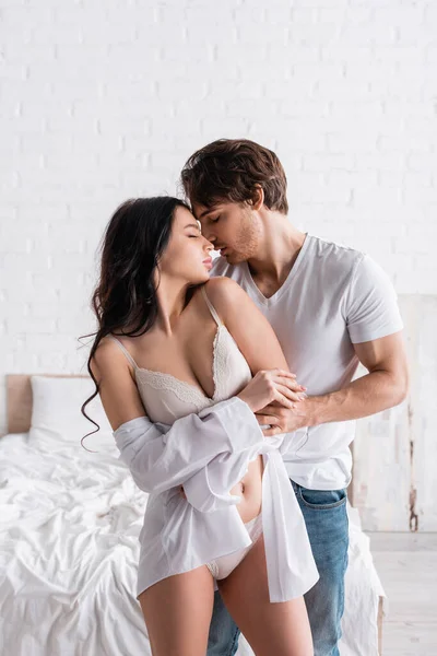 Brunette woman in white shirt and lingerie near man with closed eyes hugging her in bedroom — Stock Photo