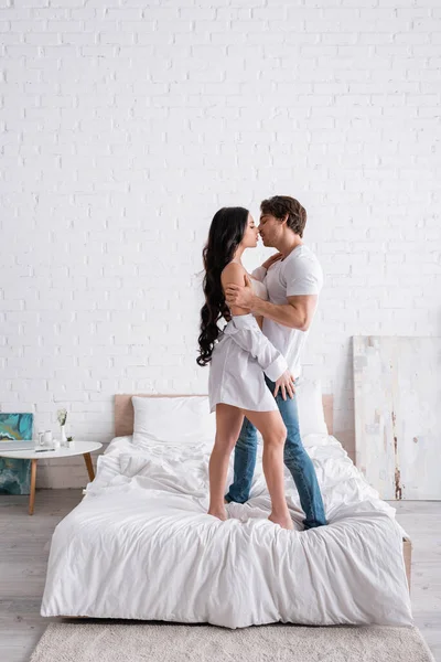 Full length view of sexy couple embracing and kissing while standing on bed — Stock Photo