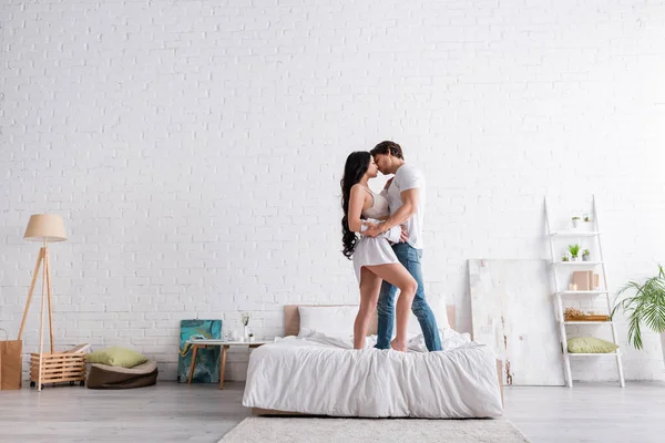 Visão de comprimento total do casal apaixonado abraçando enquanto está de pé na cama no quarto espaçoso moderno — Fotografia de Stock