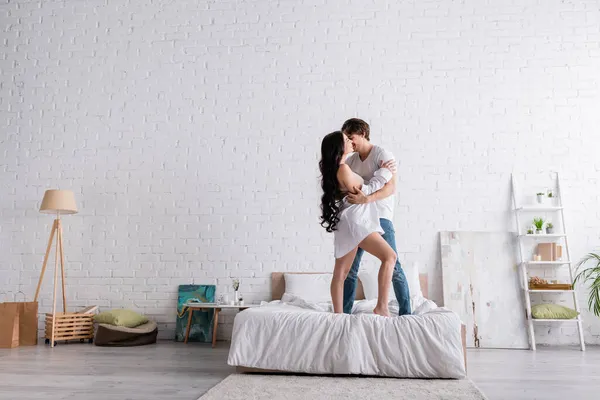 Full length view of young and sexy couple embracing while standing on bed in spacious bedroom — Stock Photo