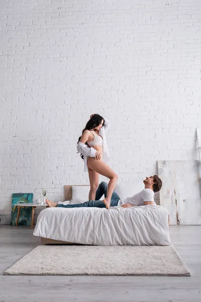 Сексуальна жінка в білій сорочці і білизні, що стоїть на ліжку біля чоловіка — стокове фото