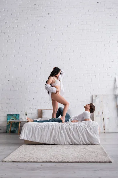 Mujer sexy en camisa blanca y lencería de pie en la cama cerca del hombre — Stock Photo