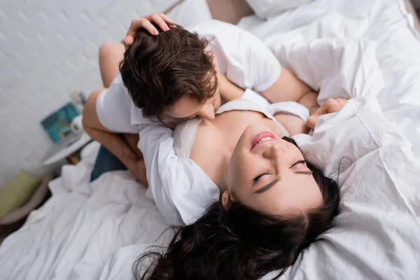 Happy and sexy woman in bra lying on bed while boyfriend kissing her body — Stock Photo