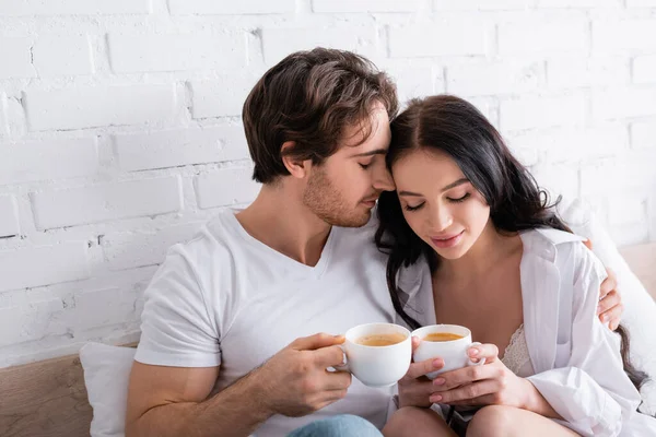 Glückliches junges Paar sitzt mit Tassen Kaffee im Bett und umarmt sich mit geschlossenen Augen — Stockfoto