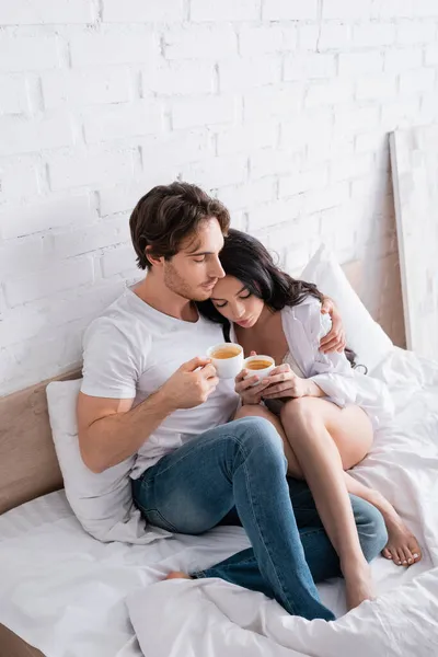 Sexy junges Paar mit Tassen Kaffee umarmt im Bett mit geschlossenen Augen — Stockfoto