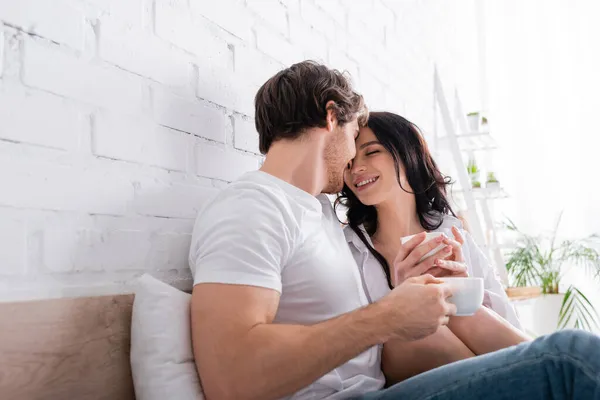 Joven pareja enamorada sonriendo mientras se sienta en la cama con tazas de café - foto de stock