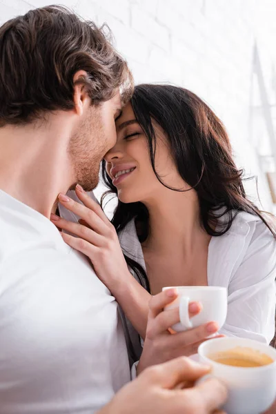 Чувственная брюнетка женщина и молодой человек, держащий кофе, обнимаясь в спальне — стоковое фото