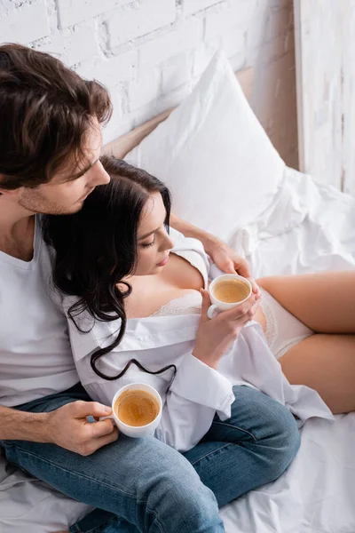 High angle view of man in jeans and sexy woman in shirt and lingerie drinking coffee on bed — Stock Photo