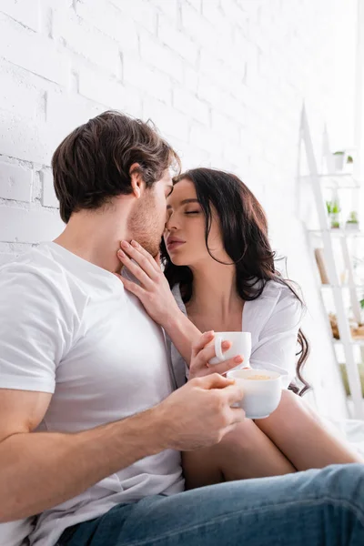 Sexy joven pareja con tazas de café besos en el dormitorio - foto de stock