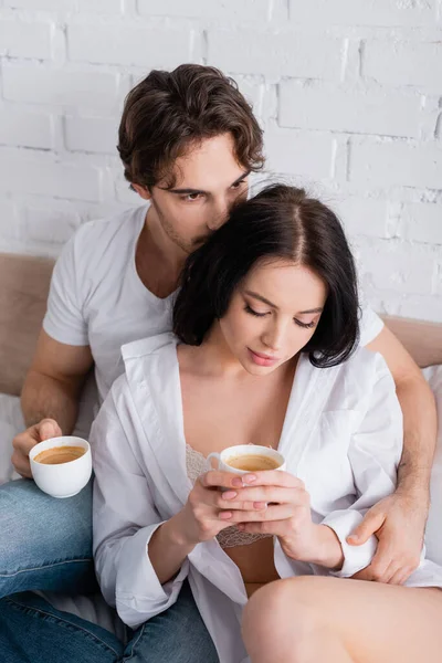Young man hugging sensual brunette woman in shirt and bra while drinking coffee on bed — Stock Photo