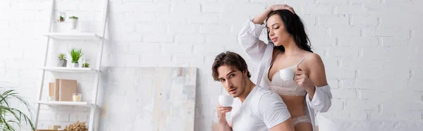 Молода і сексуальна пара тримає чашки кави в спальні, банер — стокове фото