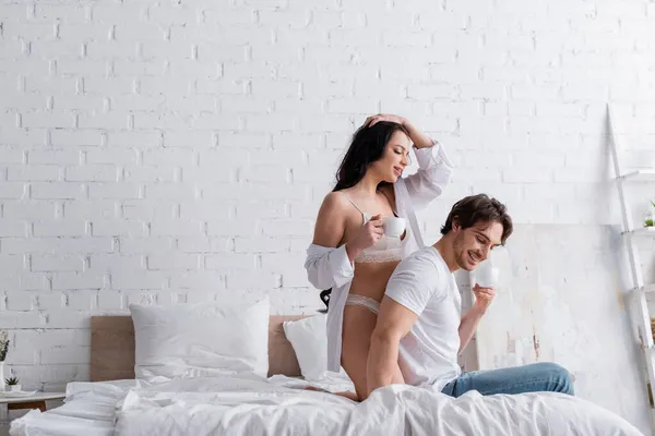 Sexy joven pareja sonriendo mientras bebiendo café de la mañana en el dormitorio - foto de stock