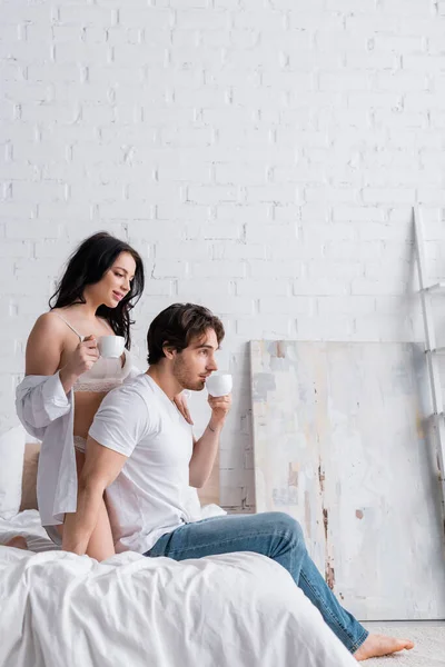 Man in jeans and seductive woman in white shirt and lingerie drinking morning coffee in bedroom — Stock Photo