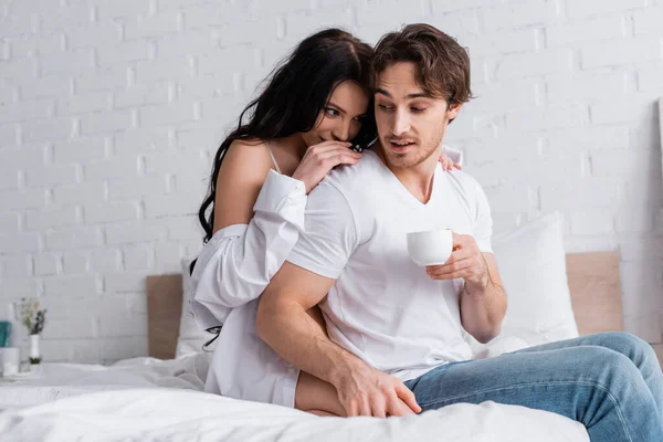 Sexy brunette woman seducing young man sitting on bed with cup of coffee — Stock Photo