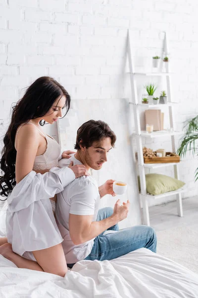 Сексуальная женщина в белой рубашке и лифчике, обнимающая плечи мужчины, пьющего кофе в спальне — стоковое фото
