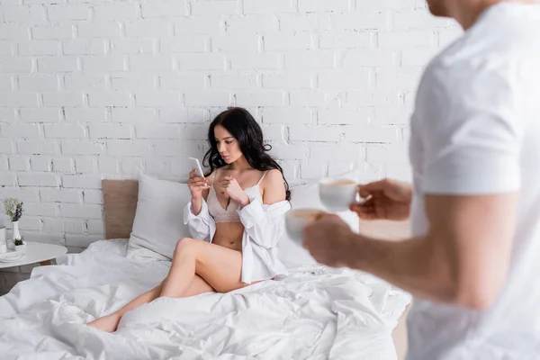 Hombre borroso sosteniendo tazas de café cerca de mujer sexy en camisa blanca y lencería usando teléfono inteligente en la cama - foto de stock