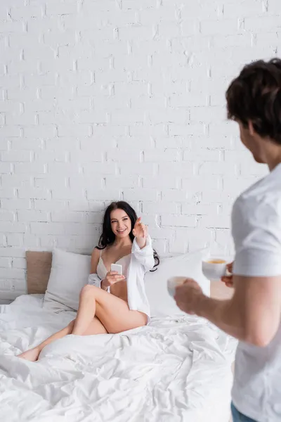 Mujer sexy y feliz con teléfono inteligente que apunta al hombre borroso con tazas de café en el dormitorio - foto de stock