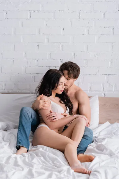 Sexy Mann in Jeans umarmt Frau in Dessous, während er mit geschlossenen Augen auf dem Bett sitzt — Stockfoto