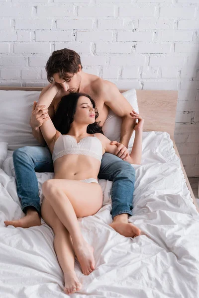 Vue grand angle de la femme passionnée en lingerie près de l'homme torse nu en jeans sur le lit — Photo de stock
