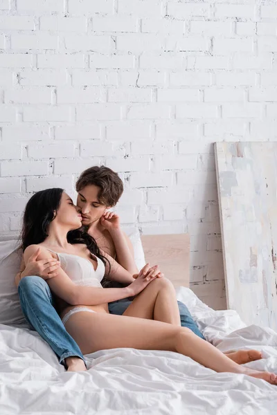 Sexy donna in biancheria intima e giovane uomo in jeans baciare sul letto vicino muro bianco — Foto stock