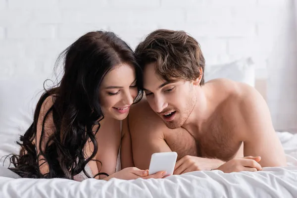 Happy and sexy couple looking at mobile phone while lying on bed — Stock Photo