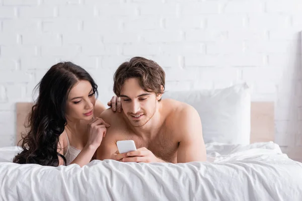 Sexy hombre sin camisa usando teléfono inteligente cerca de seductora mujer morena en el dormitorio - foto de stock