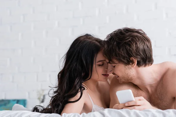 Мужчина без рубашки со смартфоном лежит на кровати рядом с сексуальной молодой девушкой — стоковое фото