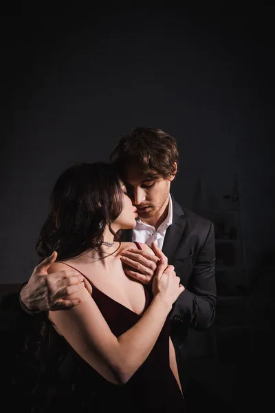 Young man in suit hugging shoulders of elegant brunette woman while seducing her in bedroom at night — Stock Photo