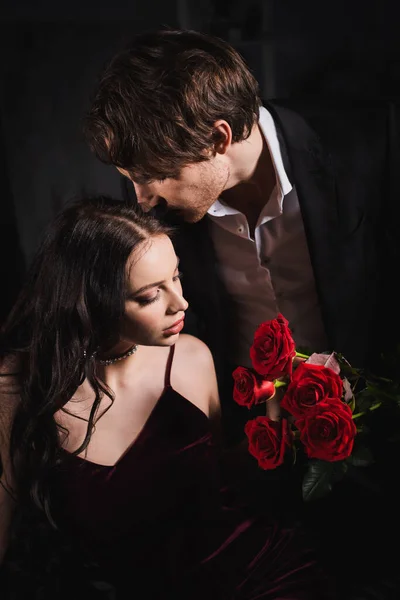 Young man holding red roses near elegant brunette woman on dark background — Stock Photo