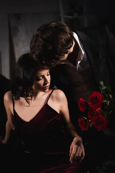 Giovane uomo che tiene rose rosse vicino a donna elegante in camera da letto scura — Foto stock