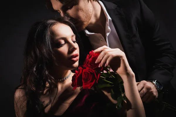 Uomo elegante con gli occhi chiusi vicino alla donna bruna che tiene rose rosse su sfondo scuro — Foto stock