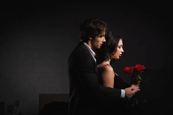 Side view of man in suit near sensual brunette woman with red roses — Stock Photo