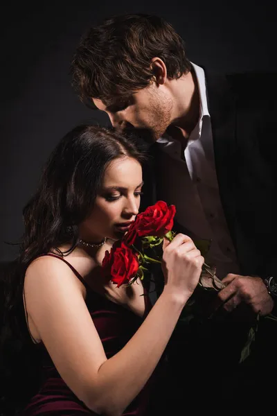 Elegante mujer oliendo rosas rojas cerca del hombre en traje sobre fondo oscuro - foto de stock