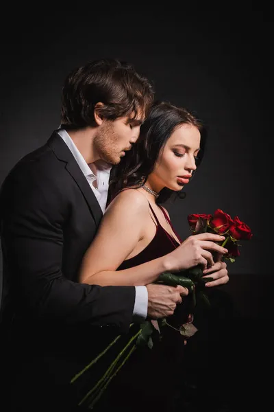 Homme aux yeux fermés embrassant femme élégante avec des roses rouges dans l'obscurité — Photo de stock