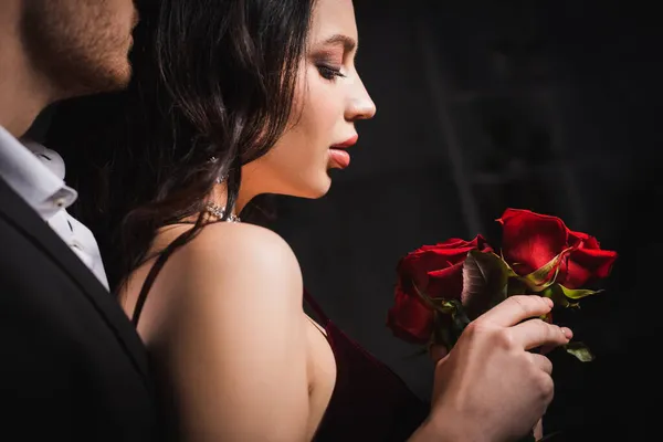 Vue latérale de la femme brune sensuelle tenant des roses rouges près du jeune homme sur fond sombre — Photo de stock