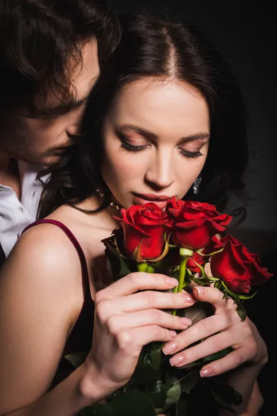 Mujer sensual y elegante sosteniendo rosas rojas cerca del hombre sobre fondo oscuro - foto de stock