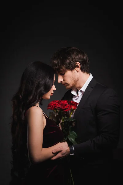 Vue latérale de la femme brune avec des roses rouges debout face à face avec l'homme sur fond sombre — Photo de stock