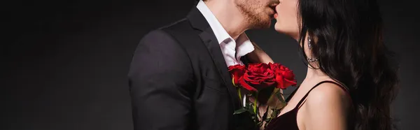 Partial view of couple in elegant clothes kissing near red roses on dark background, banner — Stock Photo