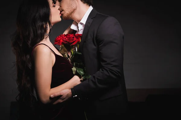 Side view of elegant young couple kissing near red roses in dark bedroom — Stock Photo