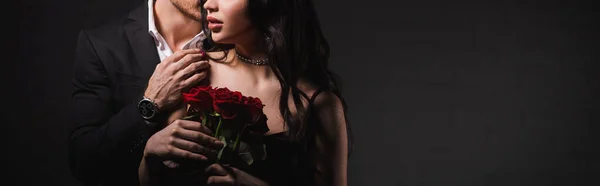 Vue recadrée de l'homme touchant l'épaule de la femme avec des roses rouges sur fond sombre, bannière — Photo de stock