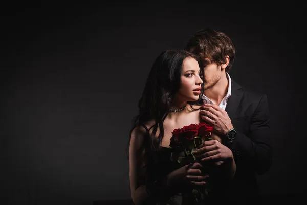Young elegant man hugging sensual brunette woman holding red roses on dark background — Stock Photo