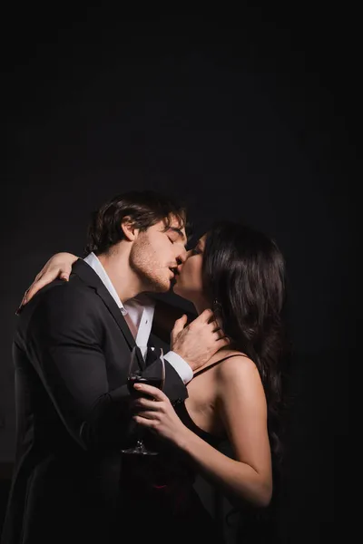 Man in black suit kissing elegant woman holding glass of red wine on dark background — Stock Photo