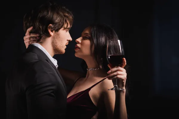 Mujer elegante con copa de vino tinto y hombre joven en traje negro mirándose el uno al otro sobre fondo oscuro - foto de stock